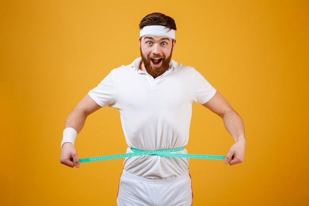 Happy bearded fitness man measuring his waist with tape